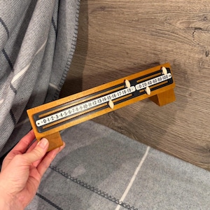 May include: A wooden counting tool featuring two horizontal tracks with sliding white markers. The bottom track displays numbers from 0 to 19, while the top track shows increments of 20, 40, 60, 80, and 100. The device has a rectangular frame with two angled support feet.