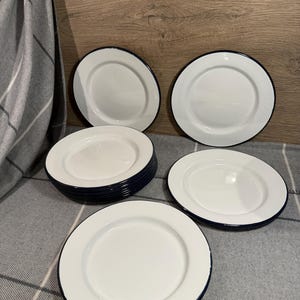 Pode incluir: Um conjunto de pratos redondos de esmalte branco com acabamento liso e uma borda azul escura distinta. A cole&ccedil;&atilde;o inclui v&aacute;rios pratos empilhados e dois pratos individuais dispostos para mostrar o design circular limpo e a superf&iacute;cie brilhante.