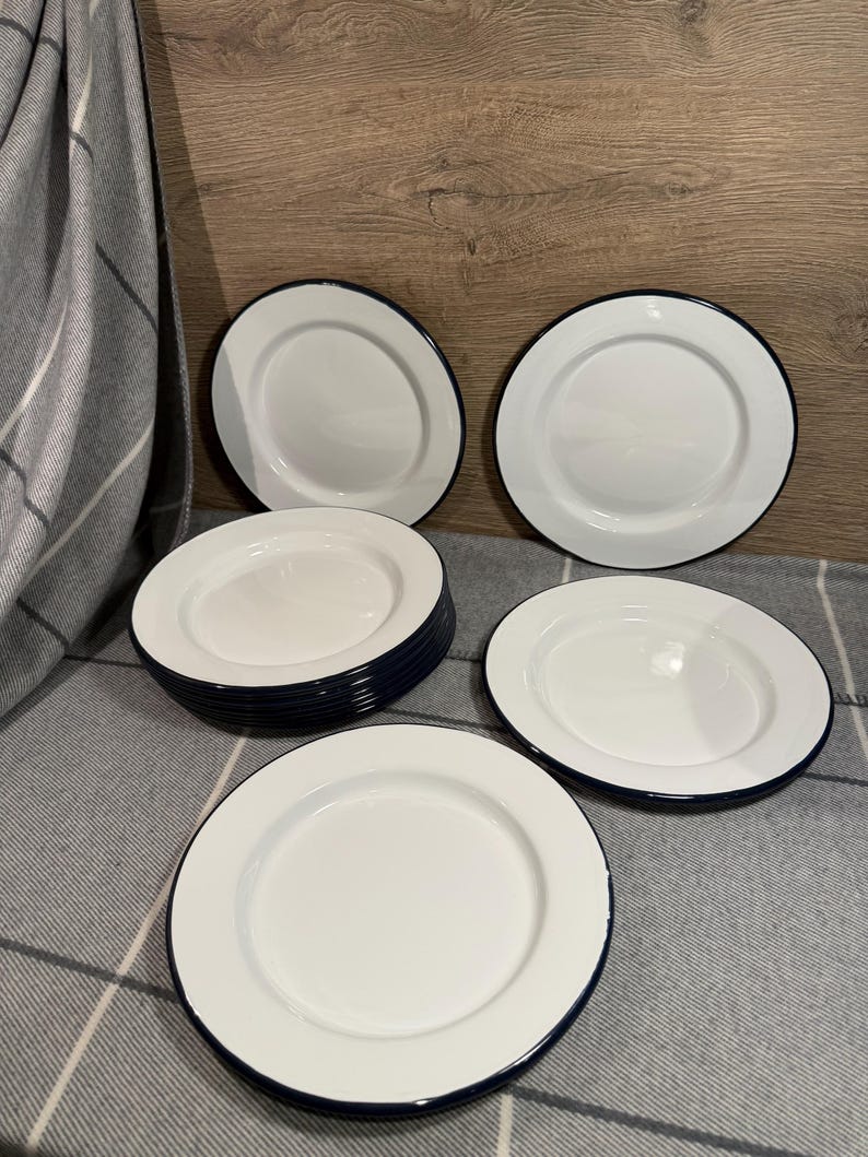 Pode incluir: Um conjunto de pratos redondos de esmalte branco com acabamento liso e uma borda azul escura distinta. A cole&ccedil;&atilde;o inclui v&aacute;rios pratos empilhados e dois pratos individuais dispostos para mostrar o design circular limpo e a superf&iacute;cie brilhante.