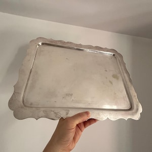 Könnte beinhalten: Ein rechteckiges Serviertablett aus Silber mit gewelltem Rand. Das Tablett wird von einer Hand vor einem weißen Hintergrund gehalten. Das Tablett hat eine glänzende, reflektierende Oberfläche.