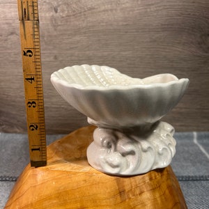 Op de afbeelding: Een witte keramische schaal in de vorm van een schelp met een golvende basis. De schaal is ongeveer 13 cm hoog.