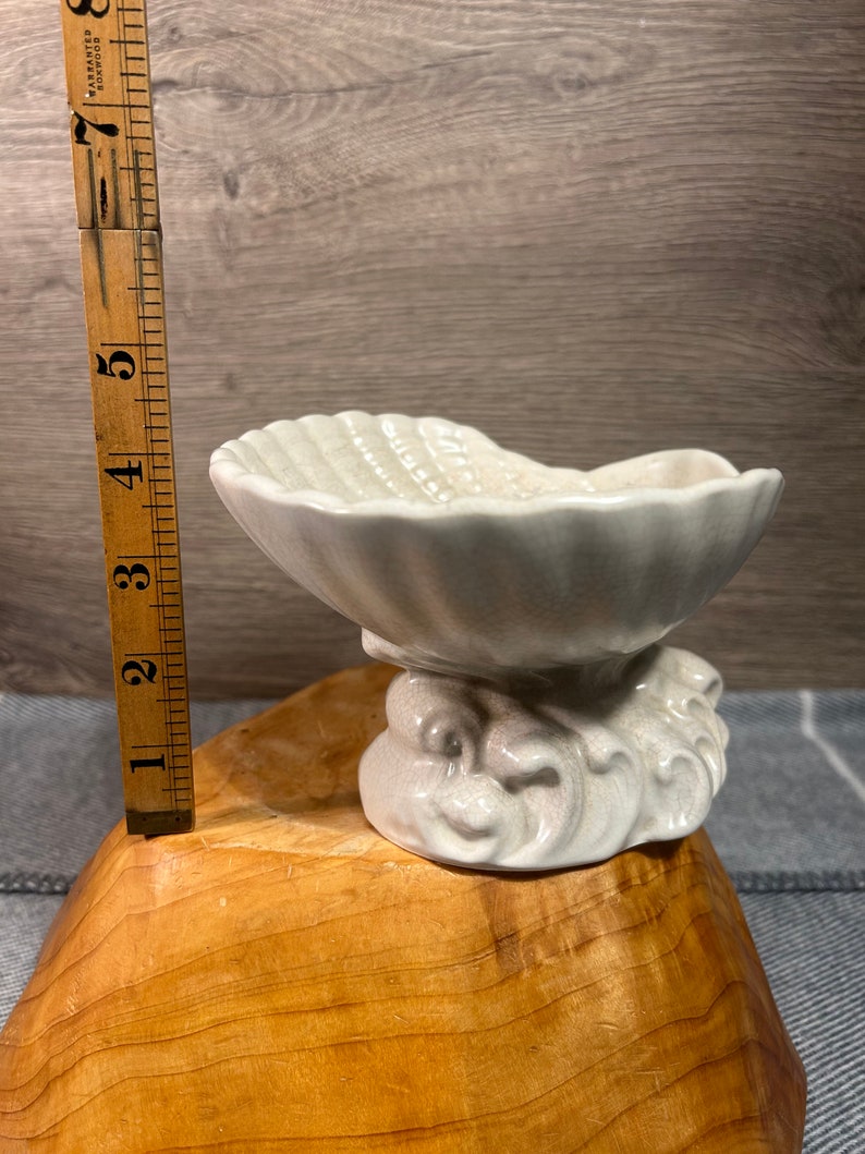 Op de afbeelding: Een witte keramische schaal in de vorm van een schelp met een golvende basis. De schaal is ongeveer 13 cm hoog.