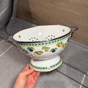 May include: A white enamel colander featuring a decorative border of green leaves and yellow lines, accented with colourful illustrations of oranges, plums, and lemons. The bowl has two silver-toned metal handles and a pedestal base with holes.