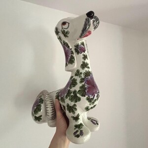 May include: A ceramic dog figurine with a white base, adorned with purple and green floral patterns. The dog has a black nose, red mouth, and is held by a person. The figurine is a decorative item.