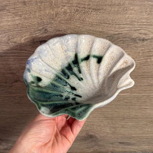 May include: A ceramic seashell-shaped dish with a white, blue, and green glaze. The dish has a scalloped edge and a textured surface. The interior features dark green and black accents. This decorative dish is ideal for holding small items.