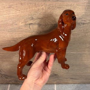 Puede incluir: Una figurita de cerámica marrón de un perro setter irlandés. El perro está de pie con la cabeza alta y la cola enroscada.
