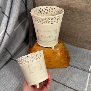 May include: Two cream-coloured ceramic flower pots with a decorative openwork rim and three small feet. The word "FLEURS" is printed in grey on the front of each pot. One pot is on a wooden base, the other is held in a hand.
