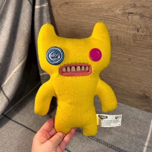 Op de afbeelding: Geel pluche speelgoed met een gestikte mond en grote tanden. Het speelgoed heeft een blauw-witte knoop voor één oog en een roze knoop voor het andere oog. Het speelgoed is gemarkeerd met "Uglydoll".