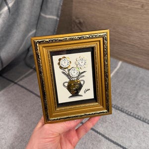 May include: A rectangular gold-toned frame with ornate detailing contains a white art print featuring a black-ink illustration of a vase holding three flowers. The flower centers are small, circular white clock faces with black Roman numerals.