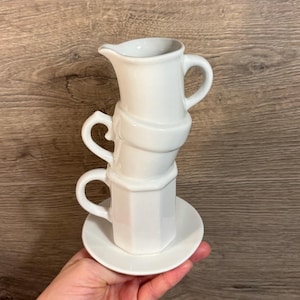 May include: A white ceramic set of stacked cups and a saucer. The cups are stacked in a unique way, with the top cup being a pitcher.