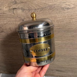 May include: A silver-toned coffee canister with a gold-colored band and lid finial. The word "COFFEE" is displayed on a gold oval plaque. The canister is held in a hand.