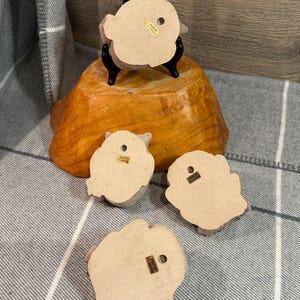 May include: Four small, light-colored owl figurines with a rough texture. One is displayed on a black stand atop a wooden base. The owls have a hole on the back for hanging. The words "Made in Taiwan" are visible on the back of each owl.