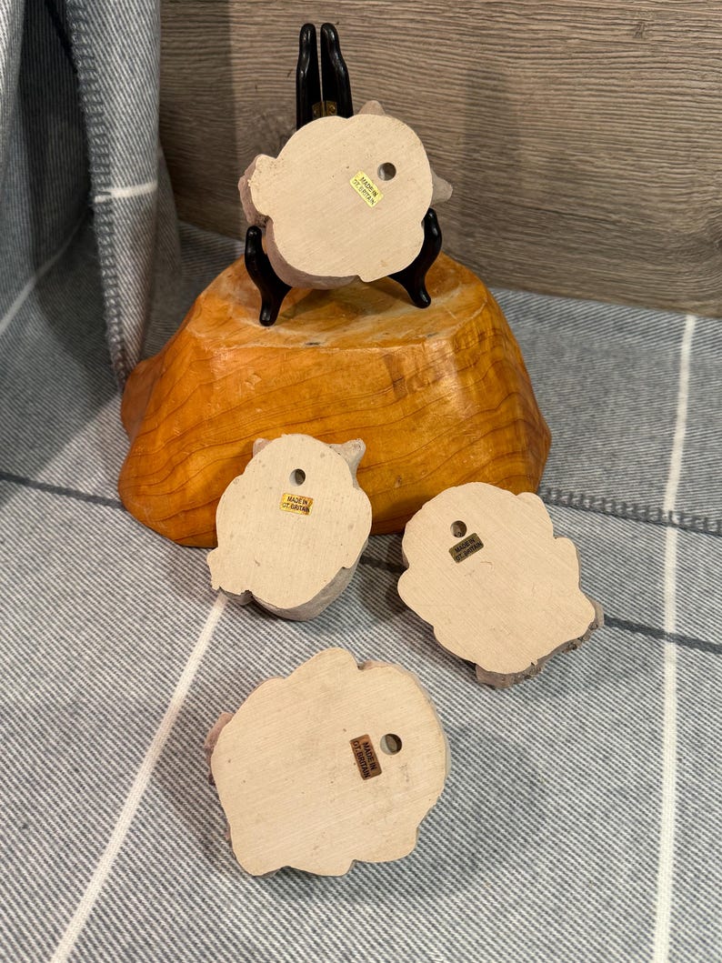 May include: Four small, light-colored owl figurines with a rough texture. One is displayed on a black stand atop a wooden base. The owls have a hole on the back for hanging. The words "Made in Taiwan" are visible on the back of each owl.