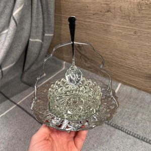 May include: A vintage clear glass butter dish with a silver-coloured metal handle and serving spoon. The dish has a decorative, starburst pattern and sits on a matching ornate tray. The spoon is suspended above the dish.