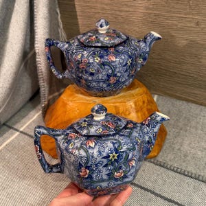 Könnte beinhalten: Zwei blaue Keramik-Teekannen mit floralen Mustern. Die Teekannen haben Deckel und Ausgüsse. Die Teekannen sind mit rosa, gelben und weißen Blumen verziert. Die Teekannen stehen auf einem Holzständer.