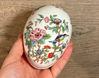 Könnte beinhalten: Eine weiße, eiförmige Schmuckschatulle aus Keramik mit einem Blumen- und Vogelmotiv. Die Schachtel ist mit rosa, blauen, grünen und roten Blumen und einem blauen Vogel mit gelbem Schnabel verziert.