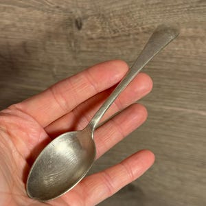 Op de afbeelding: Een zilveren lepel met een lange steel en een ovale kom. De lepel heeft een licht aangeslagen uiterlijk, wat duidt op ouderdom of gebruik. De lepel wordt in een hand gehouden tegen een houten achtergrond.