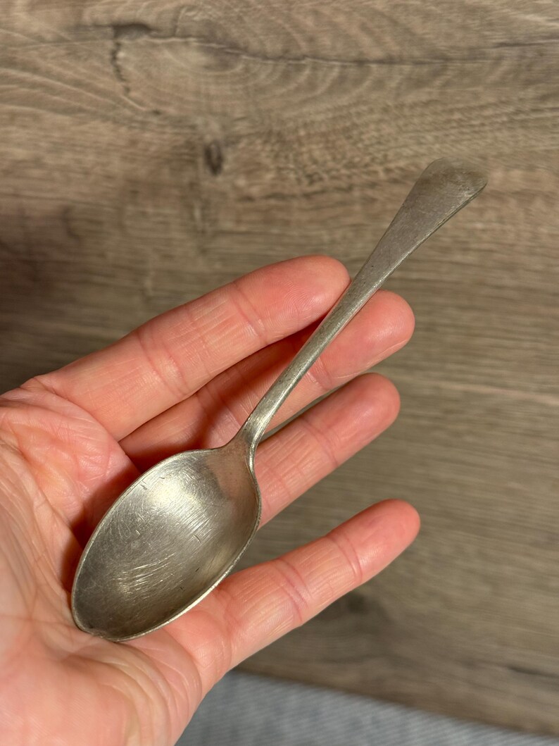 Op de afbeelding: Een zilveren lepel met een lange steel en een ovale kom. De lepel heeft een licht aangeslagen uiterlijk, wat duidt op ouderdom of gebruik. De lepel wordt in een hand gehouden tegen een houten achtergrond.