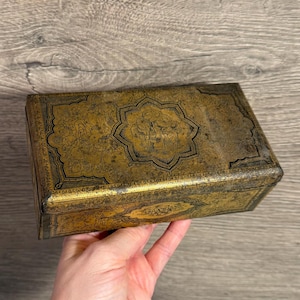 May include: A rectangular antique box with a gold and black design. The box features intricate patterns and a central floral-like motif. The box is held in a hand, showcasing its size and detail. The box appears to be made of metal.