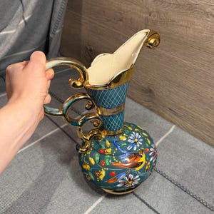 May include: A decorative ceramic pitcher featuring a teal base with a gold-trimmed handle and rim. The body is adorned with hand-painted colorful birds and floral motifs, while the neck displays a teal and gold diamond-patterned grid design.