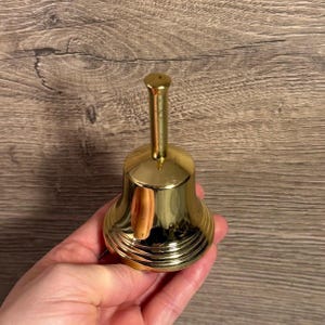 May include: A small, shiny, gold-coloured bell with a long handle. The bell has a rounded base with horizontal ridges and a smooth, polished surface. The bell is held in a hand against a wooden background.