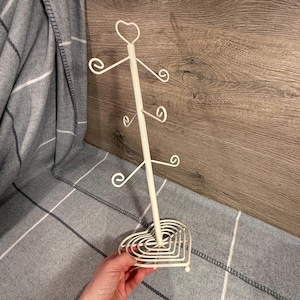 May include: A cream-coloured metal mug tree with a heart-shaped base and top. The tree has six curved arms to hold mugs and a decorative heart at the top. The mug tree is on a grey and white plaid blanket.