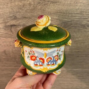May include: A green ceramic sugar bowl with a floral design and a butterfly. The lid has a yellow rose on top. The bowl has three feet.