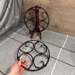May include: Two black metal trivets with a circular design. One trivet is on a wooden display stand. The other trivet is on a grey and white plaid blanket. The trivets have a decorative, swirling pattern.