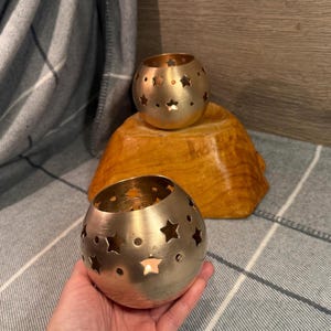 Puede incluir: Dos candelabros esféricos dorados con recortes en forma de estrella. Uno se sostiene en una mano y los otros dos están sobre una base de madera. Los candelabros están sobre una manta a cuadros gris y blanca.