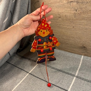 Puede incluir: Un payaso de madera de cuerda con lunares rojos y blancos, una camisa a rayas arcoíris y un gato rojo en el pecho. El payaso tiene una nariz roja y botones rojos.