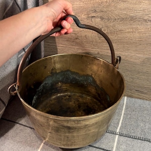 Puede incluir: Un caldero de latón antiguo con un asa curva de metal oxidado. El interior muestra signos de uso con residuos oscuros. El caldero es sostenido por una mano, lo que sugiere su tamaño y portabilidad. La estética general es rústica y antigua.