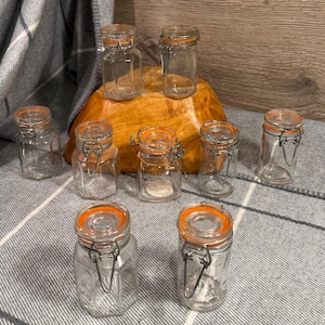 May include: A selection of clear glass jars with orange rubber seals and metal clasps. The jars vary in size and shape, some hexagonal. The jars are displayed on a wooden bowl and a grey and white striped fabric.