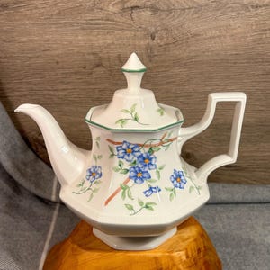 May include: White teapot with a floral design featuring blue flowers and green leaves. The teapot has a lid with a pointed top and a handle on the side.