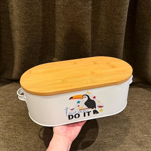 May include: An oval white metal storage bin with a light brown bamboo lid and two small side handles. The front features a graphic of a toucan perched on a branch with colorful stars and the text "toucan DO IT" printed in black and blue.