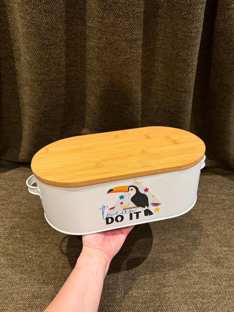 May include: An oval white metal storage bin with a light brown bamboo lid and two small side handles. The front features a graphic of a toucan perched on a branch with colorful stars and the text "toucan DO IT" printed in black and blue.
