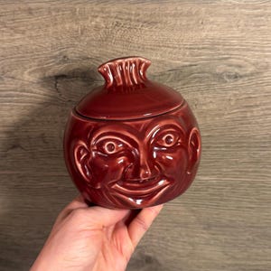 May include: A dark red ceramic jar with a lid, featuring a sculpted human face on the front. The lid has a pleated, fan-like handle at the top. The surface of the jar is glossy and smooth, reflecting light across the facial features.