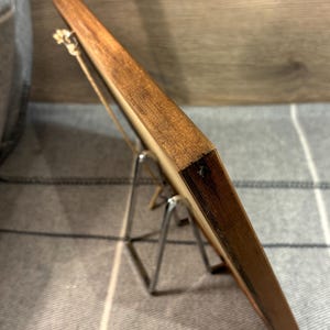 May include: A wooden picture frame with a natural wood grain finish. The frame is supported by a silver metal easel stand. The frame is rectangular and appears to be empty, ready for a photo or artwork.