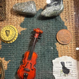 Puede incluir: Un collage enmarcado de recuerdos irlandeses, que incluye un viol&iacute;n en miniatura, una tapa de botella de Guinness, una moneda de 2 peniques, un sello de correos con un mirlo y dos piedras verdes.