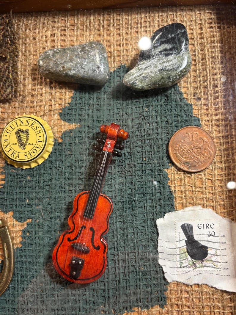 Puede incluir: Un collage enmarcado de recuerdos irlandeses, que incluye un viol&iacute;n en miniatura, una tapa de botella de Guinness, una moneda de 2 peniques, un sello de correos con un mirlo y dos piedras verdes.