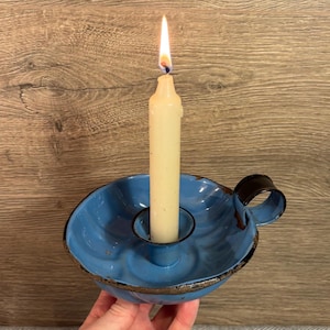 May include: A blue metal candle holder with a handle. The holder has a scalloped edge and a worn, rustic look. A white candle is burning in the holder.
