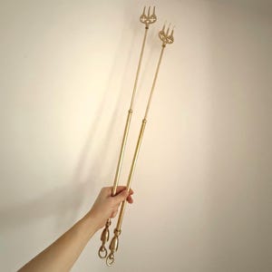 May include: Two gold-coloured fireplace tools with decorative fork-shaped tops and ringed handles. The tools are held against a white background. The tools are likely used for tending a fire.