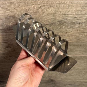 Puede incluir: Un tostador de acero inoxidable con base rectangular y separadores arqueados. El soporte está diseñado para sostener varias rebanadas de tostadas en posición vertical. El metal tiene una superficie brillante y reflectante.