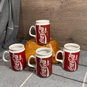 May include: Four ceramic Coca-Cola mugs. Each mug is red with white accents and features the Coca-Cola logo and the word "Coke". The mugs are arranged on a wooden surface and a patterned cloth.
