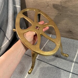 May include: A brass-coloured metal trivet with a circular design and three curved legs. The trivet has a wheel-like appearance with open spaces. The object is held by a person's hand.