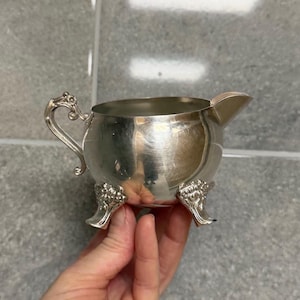 Può includere: Una piccola lattiera d'argento con corpo arrotondato, beccuccio e manico decorativo. La lattiera poggia su tre piedini ornati. La superficie argentata riflette la luce, conferendole un aspetto lucido. Un pezzo classico per servire panna o latte.