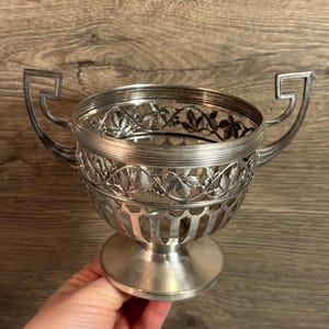 Pode incluir: Tigela decorativa prateada com duas alças. A tigela apresenta um design detalhado de folhas e videiras ao redor da borda e uma base de pedestal. A tigela tem um design recortado e provavelmente é feita de metal.