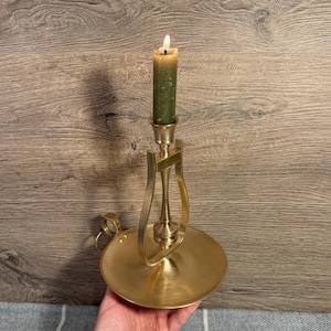 May include: A brass candlestick with a lit green candle. The candlestick has a round base, a curved handle, and a decorative support for the candle. The candle is cylindrical and has a visible flame.