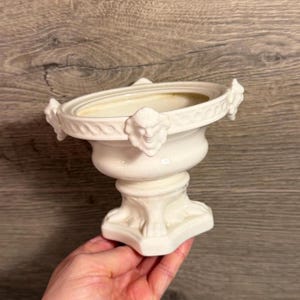 Pode incluir: Uma urna de cerâmica na cor creme com uma borda circular larga decorada com motivos de rostos humanos esculpidos. O recipiente tem um corpo alargado apoiado numa base de pedestal com pés em forma de garra e um acabamento liso e brilhante.