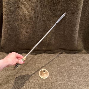 Puede incluir: Una varilla metálica larga y delgada con una punta afilada y un mango texturizado de color latón, acompañada de una pequeña base circular separada de color latón con un orificio central. La varilla tiene un acabado metálico.