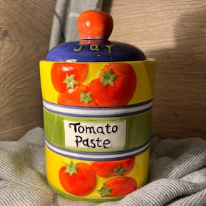 May include: A ceramic tomato paste jar with a lid. The jar is yellow with red tomato illustrations and green accents. The lid is blue with the word "jar" and a tomato-shaped knob. The jar has the words "Tomato Paste" on the front.